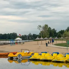 Żółte rowerki wodne i łódki czekają na plaży nad jeziorem blisko Warszawy. Para spaceruje po piasku pod parasolami.