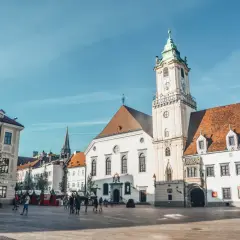 Słoneczny dzień na bratysławskim rynku. Ludzie spacerują po brukowanym placu przed zabytkowymi budynkami z charakterystyczną wieżą zegarową.