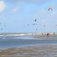 Wietrzny dzień na plaży w Holandii. Kolorowe latawce do kitesurfingu unoszą się nad falującym morzem, a ludzie spacerują po mokrym piasku.