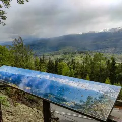 Widok z wieży widokowej Karkonosze. Na pierwszym planie tablica informacyjna z mapą panoramy, w tle góry i lasy.