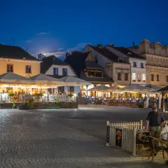Wieczorny rynek w Kazimierzu Dolnym tętni życiem. Ludzie siedzą przy stolikach pod parasolami, ciesząc się letnim wieczorem.