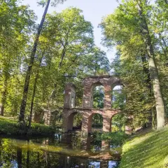 Kamienne ruiny mostu w Arkadii Nieborów odbijają się w spokojnej wodzie, otoczone bujną zielenią drzew.