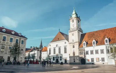 Słoneczny dzień na bratysławskim rynku. Ludzie spacerują po brukowanym placu przed zabytkowymi budynkami z charakterystyczną wieżą zegarową.
