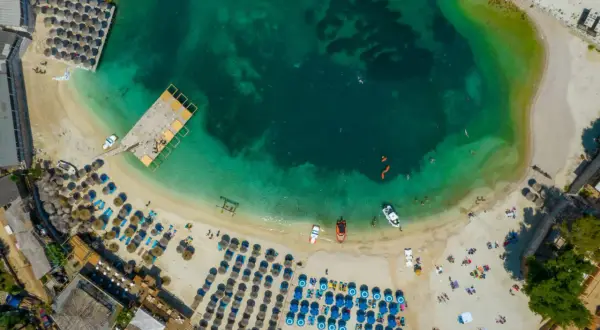 Widok z lotu ptaka na plażę z rzędami parasoli i leżaków, turkusową wodą i łodziami. Idealne miejsce na wakacje w albańskich miastach nad morzem.