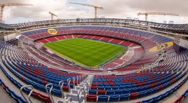 Camp Nou w Barcelonie w trakcie modernizacji. Widok na pusty stadion z zieloną murawą i czerwono-niebieskimi trybunami, nad którymi górują dźwigi budowlane.