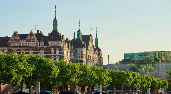 Kopenhaga ceny: zabytkowe budynki i nowoczesna architektura nad wodą, z rzędem drzew i zaparkowanymi samochodami.