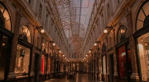 Elegancka galeria handlowa w Brukseli, z rzędem sklepów i kawiarni, ozdobiona złotymi, wiszącymi instalacjami. To urokliwe miejsce, które warto odwiedzić, zwiedzając brukselskie dzielnice.