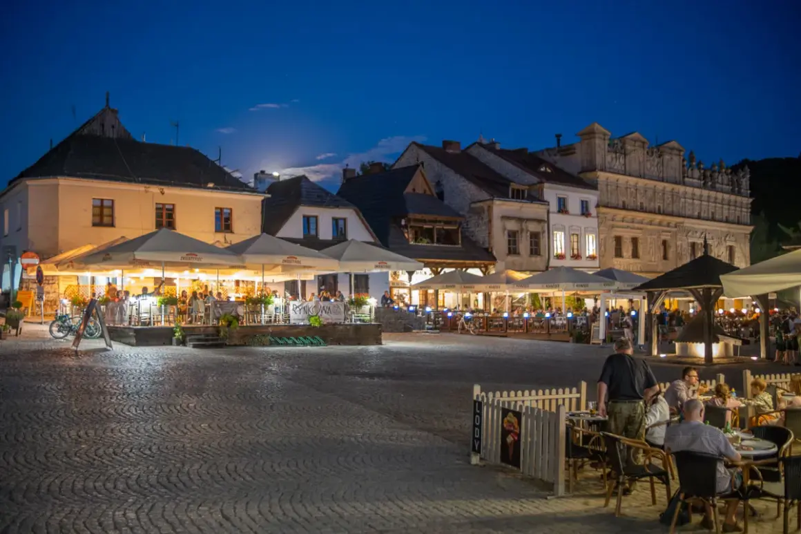 Wieczorny rynek w Kazimierzu Dolnym tętni życiem. Ludzie siedzą przy stolikach pod parasolami, ciesząc się letnim wieczorem.
