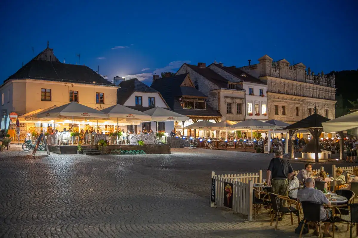 Wieczorny rynek w Kazimierzu Dolnym tętni życiem. Ludzie siedzą przy stolikach pod parasolami, ciesząc się atmosferą.