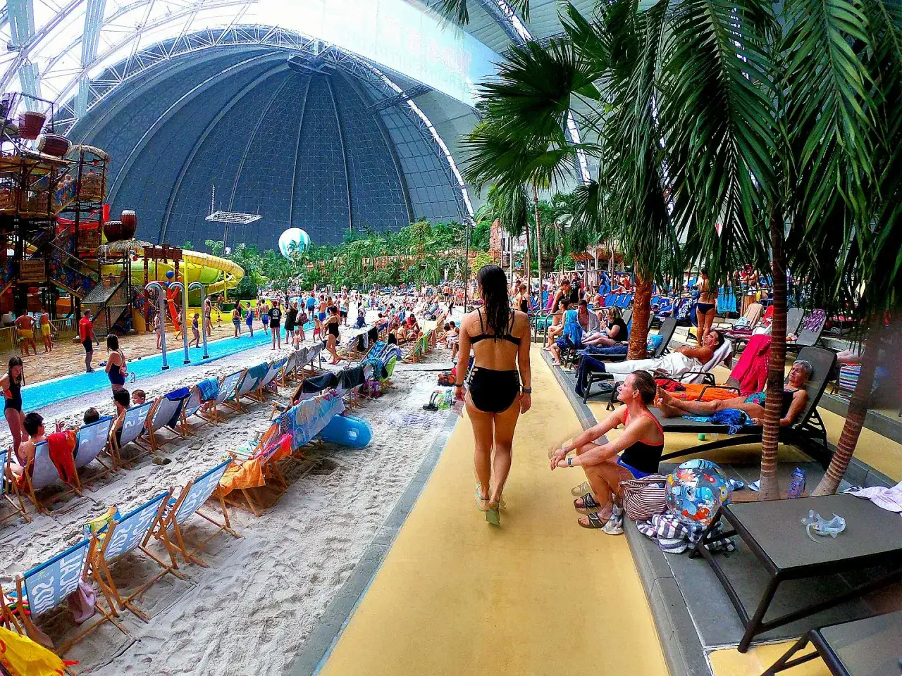 Tropikalny aquapark pod Berlinem z palmami, basenami i zjeżdżalniami. Ludzie odpoczywają na leżakach i spacerują po piaszczystej plaży.