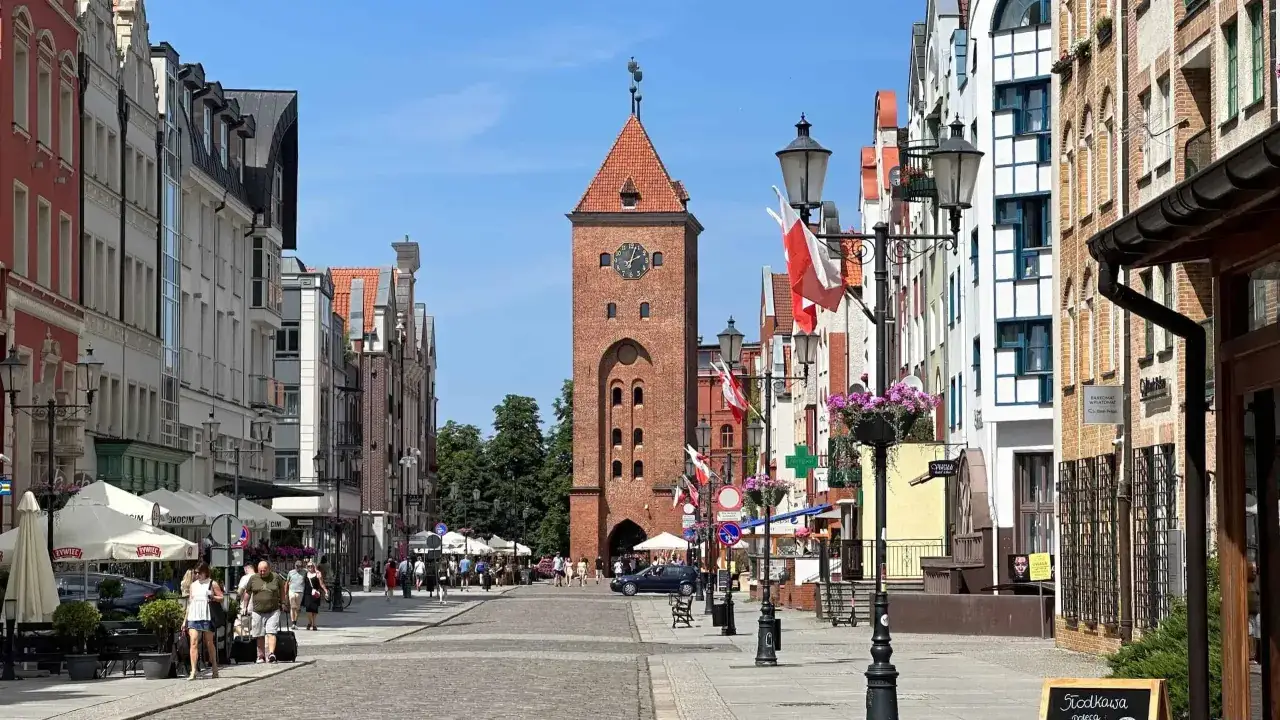 Spacer po uliczkach starego miasta Elbląg. W oddali widać charakterystyczną wieżę z zegarem, a po bokach kolorowe kamienice i kawiarniane ogródki.