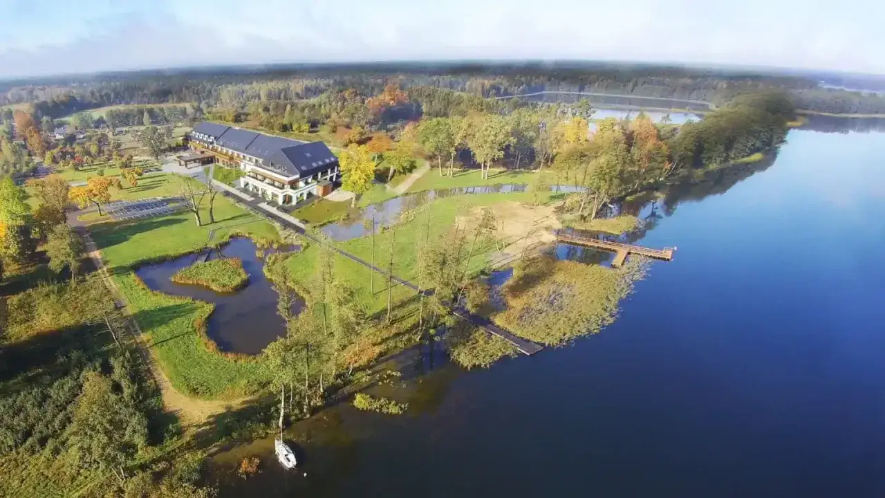 Malownicze mazury miejscowości nad jeziorem. Hotel z pomostem i łódką przy spokojnej tafli wody, otoczony zielenią i jesiennymi drzewami.