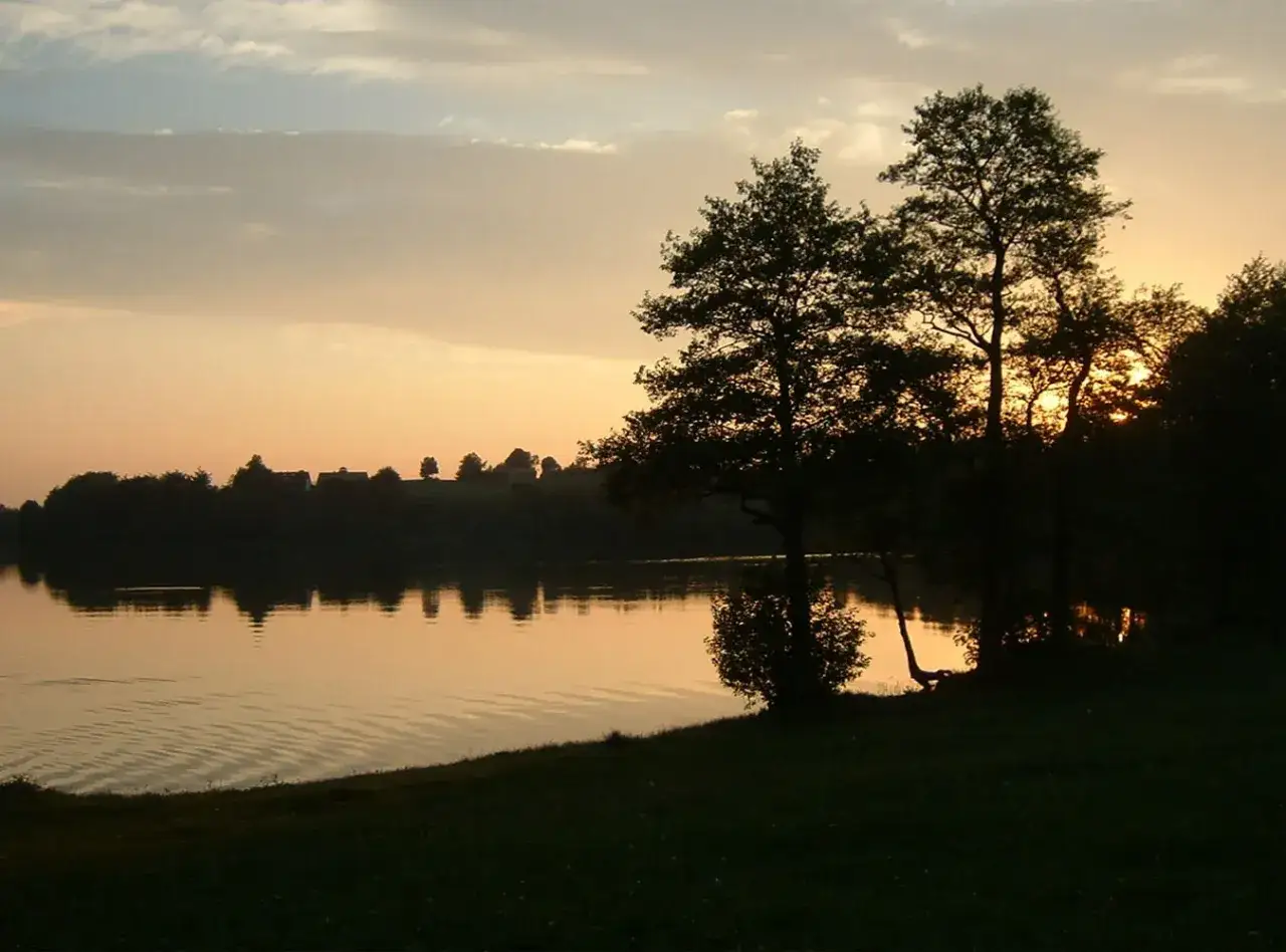 Zachód słońca nad jeziorem, z sylwetkami drzew i domów na wzgórzu. Widok przypomina sielankowe szwajcarskie kaszuby.