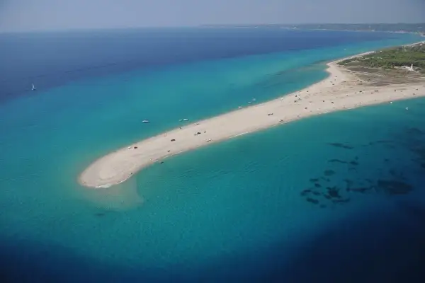 Plaża na Chalkidiki, przypominająca język lądu wcinający się w turkusowe morze. W oddali widać wybrzeże i budynki, może nawet lotnisko.