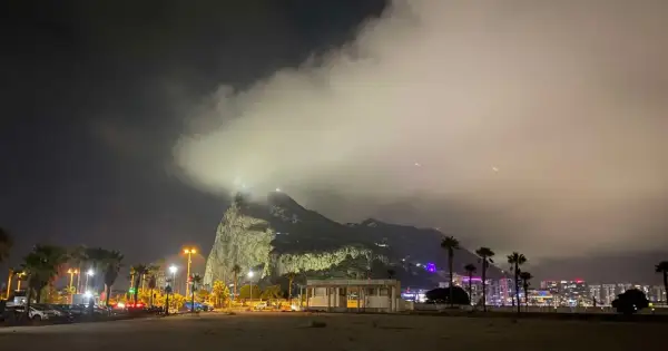 Nocny widok na Gibraltar, z chmurami otulającymi skałę. W oddali widać oświetlone miasto i palmy.