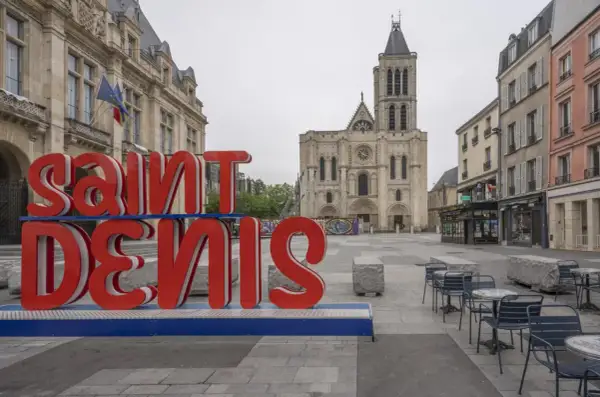 Duże czerwone litery "SAINT DENIS" na tle bazyliki. Choć nazwa sugeruje niebezpieczne miejsca, plac wydaje się spokojny.