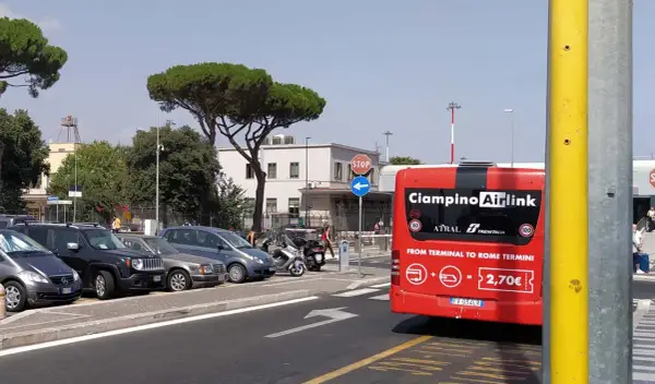 Czerwony autobus Ciampino Airlink czeka na pasażerów. Z tyłu widnieje napis "FROM TERMINAL TO ROME TERMINI" i cena 2,70€. W tle parking z samochodami i budynek lotniska.